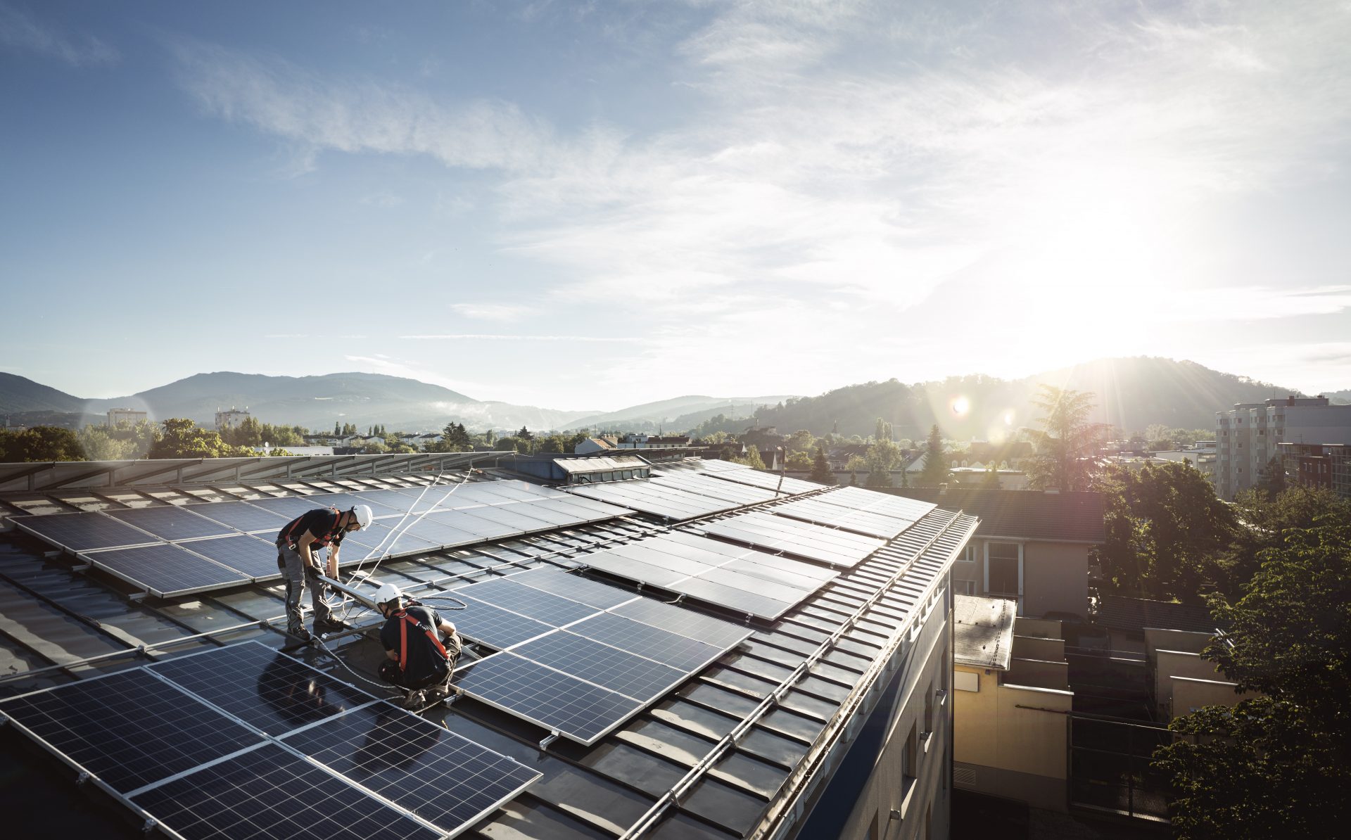 Zwei E-Werk Franz Mitarbeiter bei Montagearbeiten einer Photovoltaik-Anlage auf einem Dach mit Blick auf die Göstinger Umgebung bei Sonnenlicht